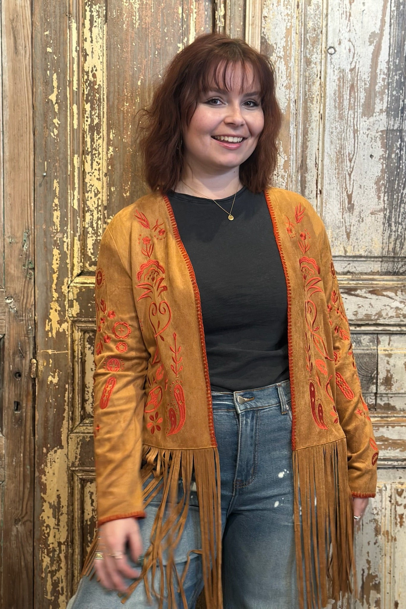 Woman wearing a brown embroidered cardigan with fringe in front of a rustic wooden door.